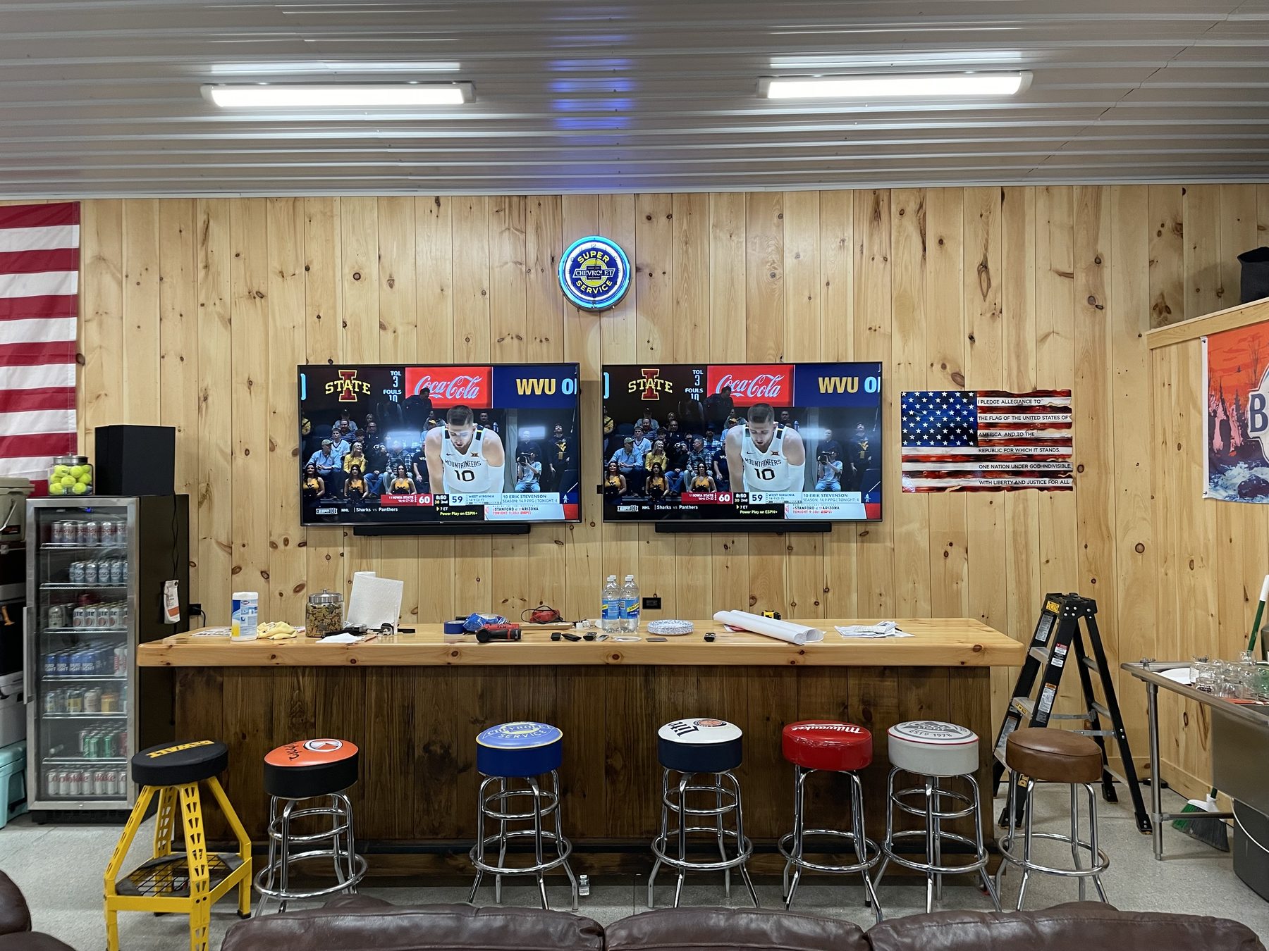 Two 75-inch flat-panel TVs mounted side by side on a knotty-pine plank wall in a home sports bar, both showing an Iowa State basketball game, with dual soundbars below the panels and four surround speakers placed around the room, polished wood bar, American flag and team-logo decor, and pendant industrial lighting overhead
