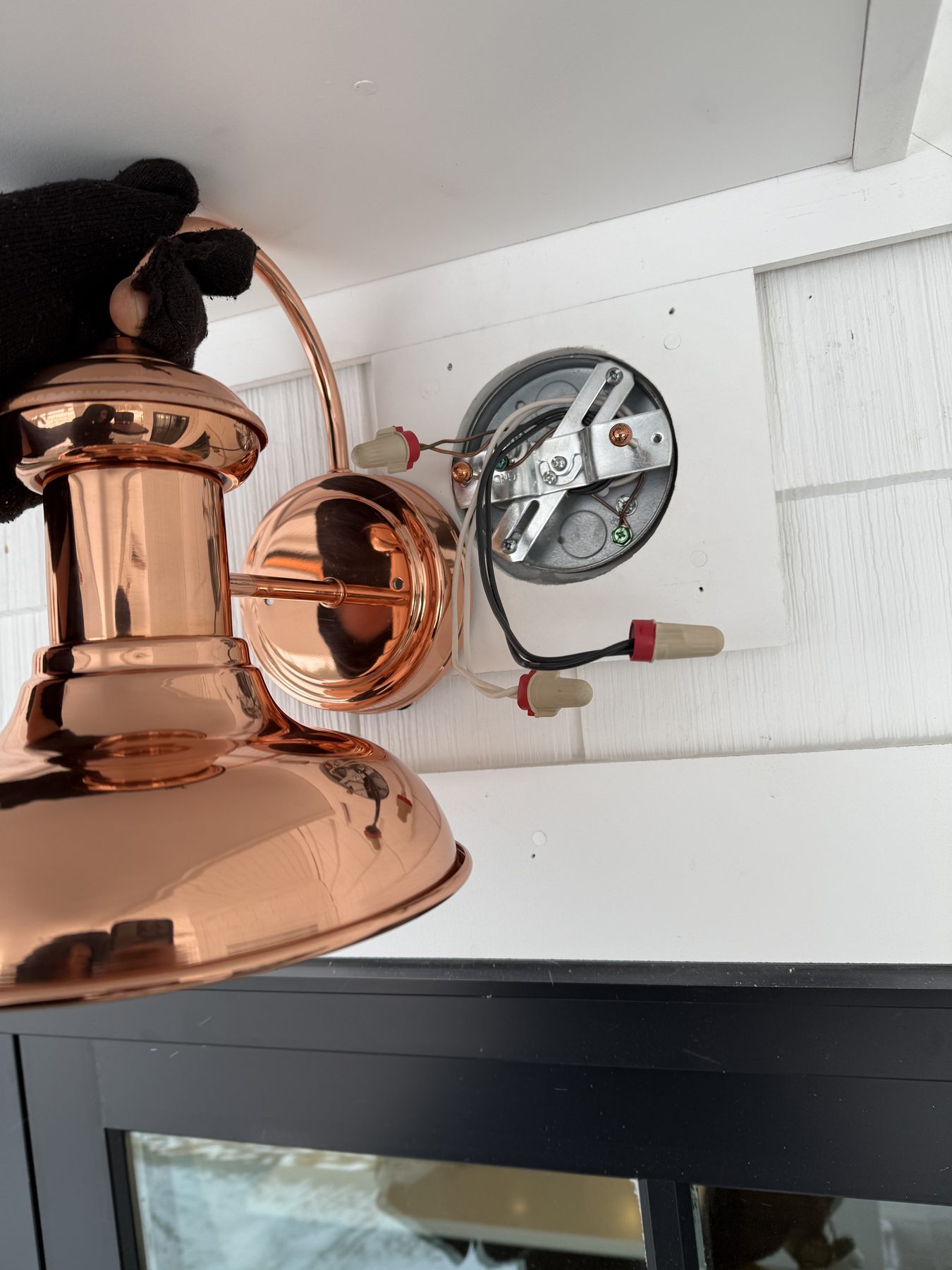 Polished copper wall sconce being installed against white siding, gloved hand visible holding the fixture, junction box and wire nuts in frame