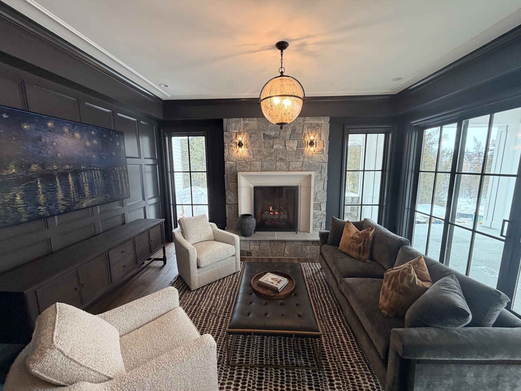 Wide view of the same living room from a different angle, showing the TV in art mode displaying Van Gogh's Starry Night as the artwork background against the dark coffered wall, gray sofa, ottoman, stone fireplace with paired sconces, and a gold sphere chandelier suspended overhead