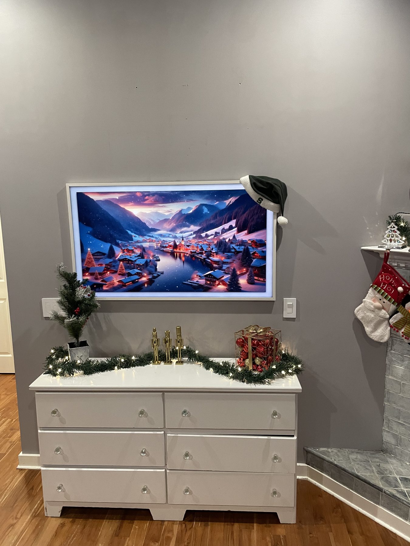 Wall-mounted Samsung Frame TV in art mode showing a stylized alpine village painting, with holiday garland and a Santa hat hanging from the corner