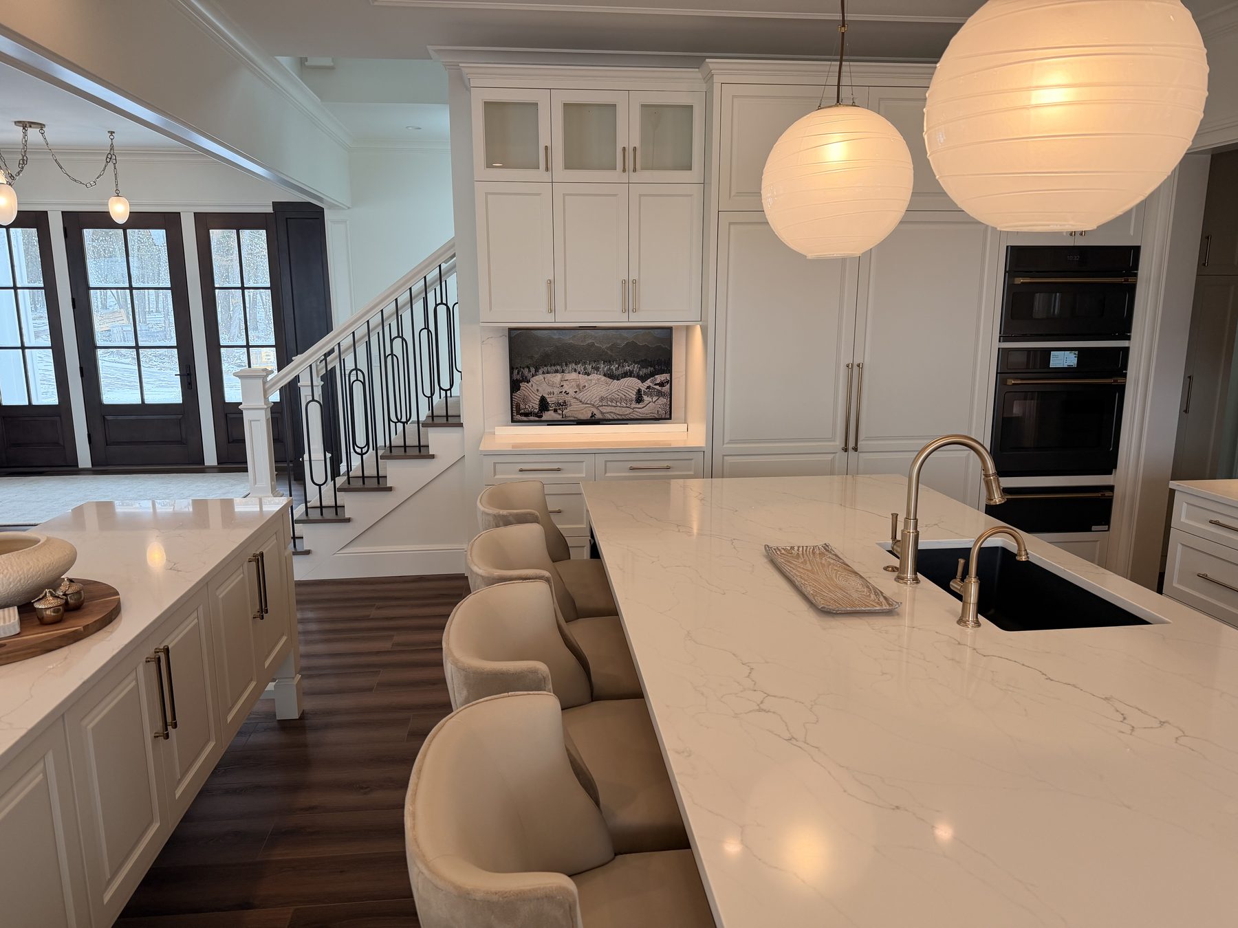Bright white kitchen with quartz island, gold faucet, ribbed glass globe pendant fixtures, a motorized TV mounted into the cabinetry that drops down from a hidden bay behind the upper cabinets, and a built-in double oven