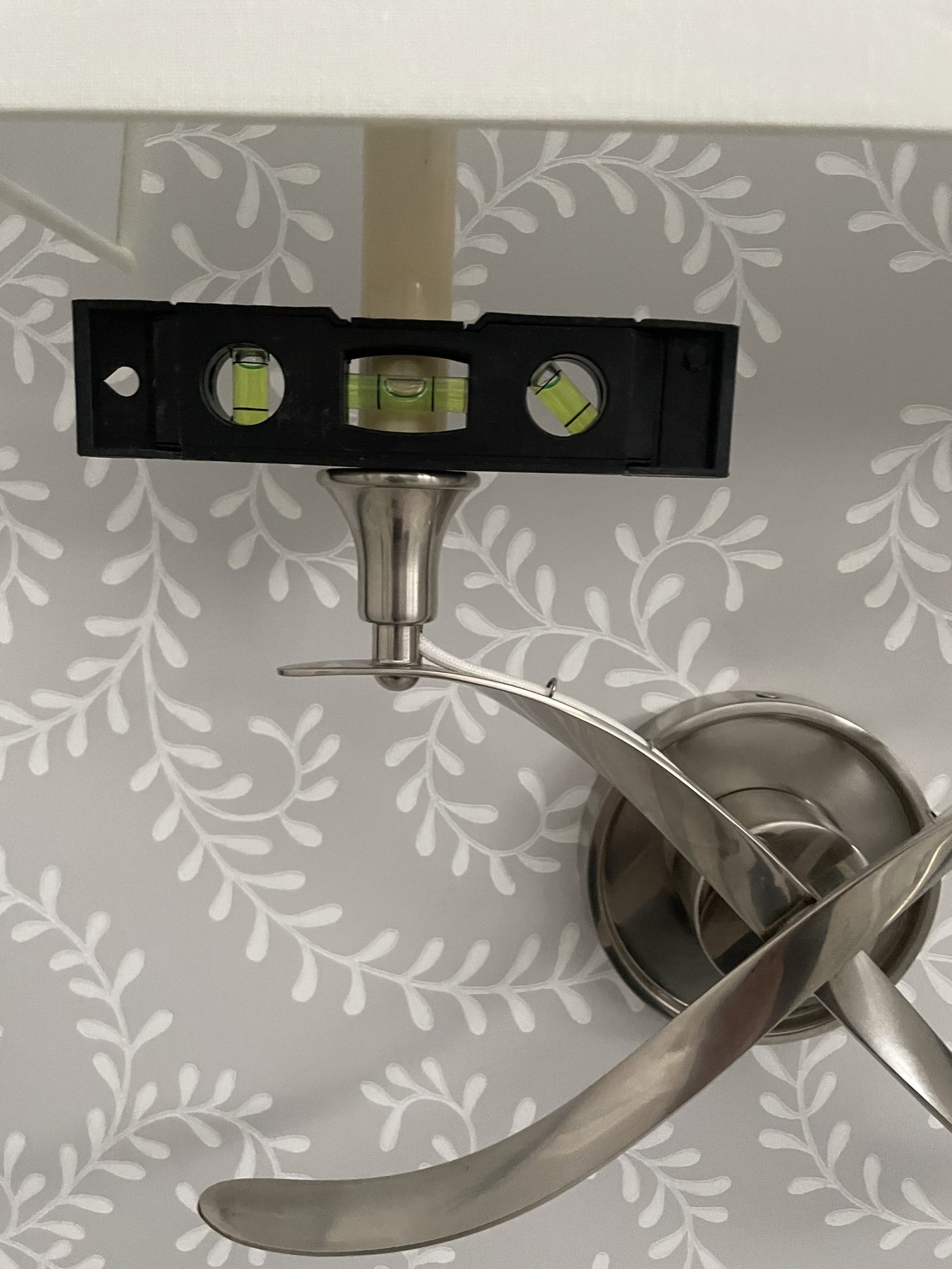Torpedo level held against the horizontal arm of a brushed-nickel candelabra-style wall sconce on botanical wallpaper, bubble centered confirming the fixture is perfectly level