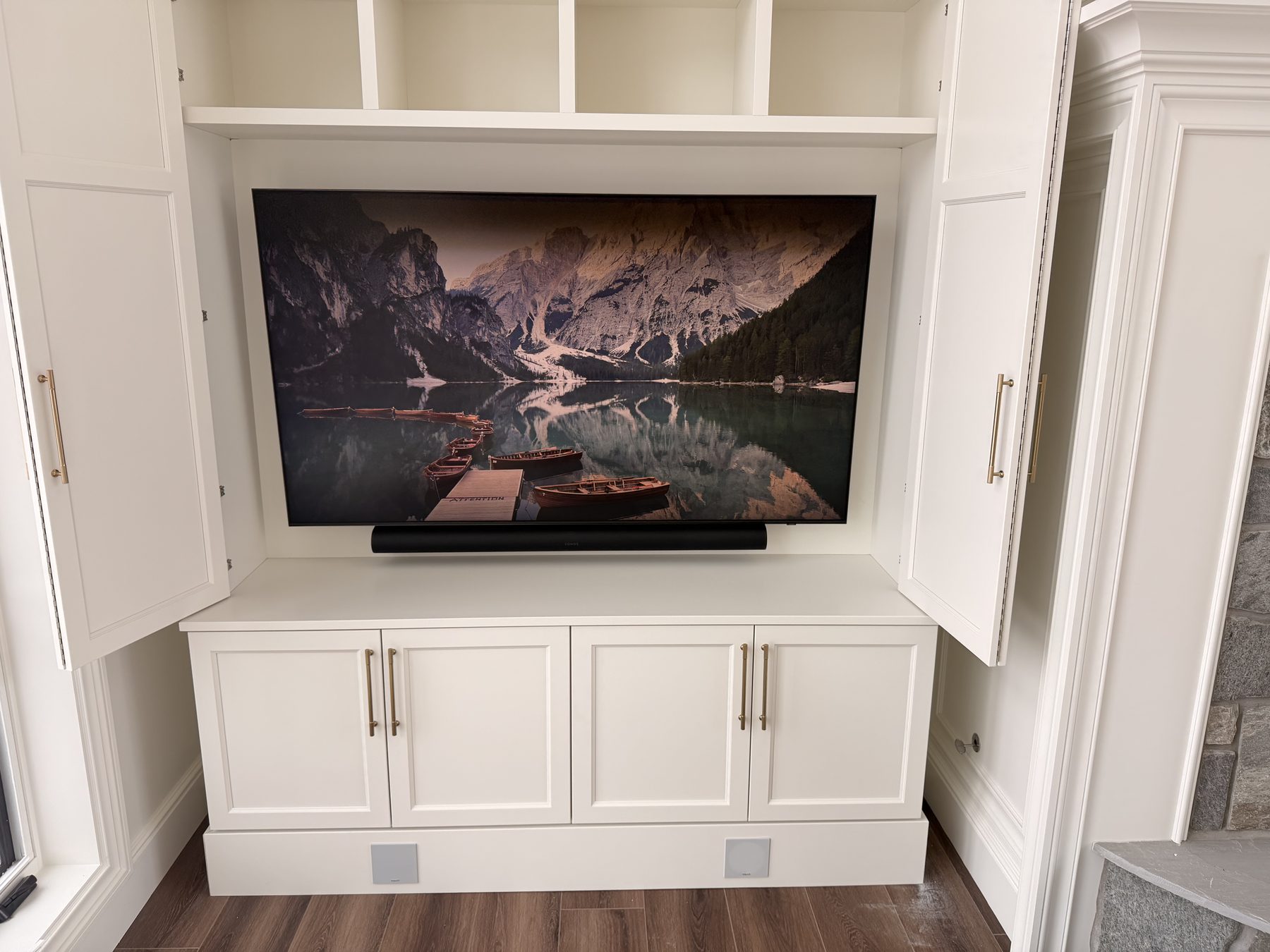 Large TV showing a mountain lake landscape installed in a white painted millwork cabinet with a soundbar below, and two black speaker grilles in the kickboard concealing a pair of Klipsch ported subwoofers