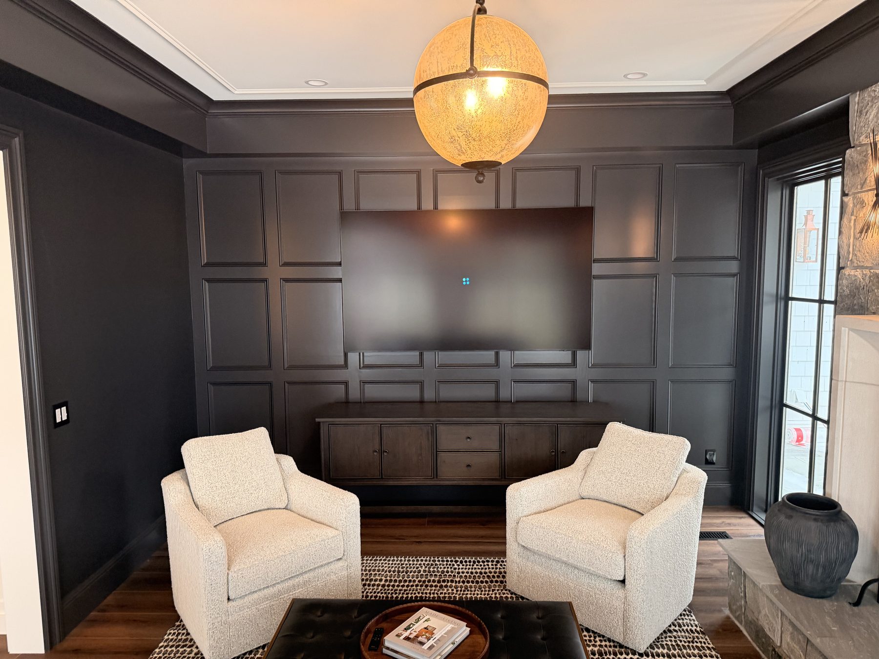 Large flat-panel TV mounted inside a dark coffered millwork wall in a formal den, with two cream boucle armchairs facing it, a spherical pendant fixture overhead, and an in-floor subwoofer hidden in the floor behind the right-hand chair