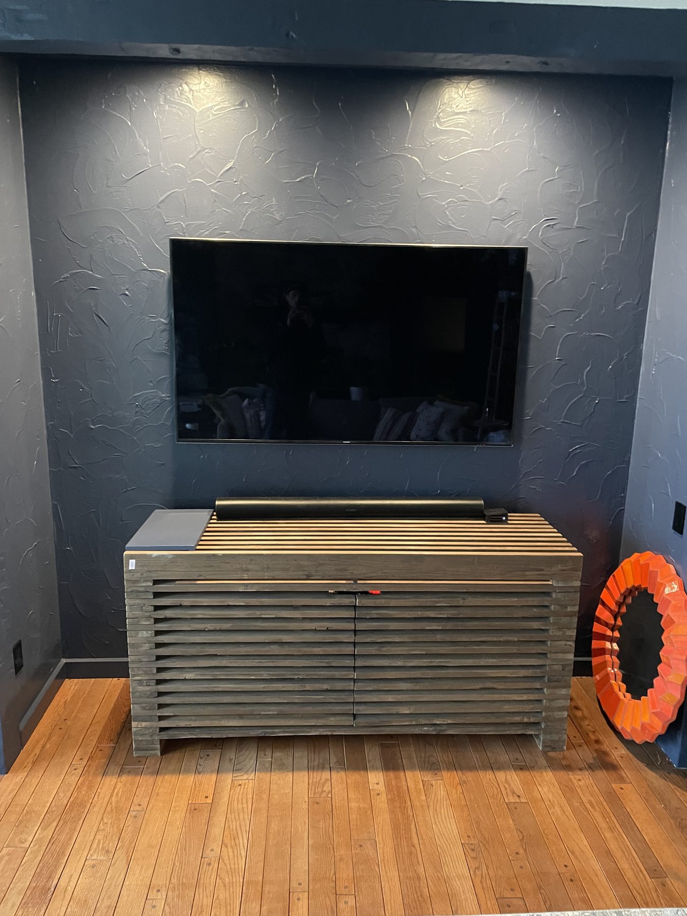Large flat-panel TV wall-mounted on a textured charcoal plaster wall above a horizontal wood-slat A/V cabinet with downlights above