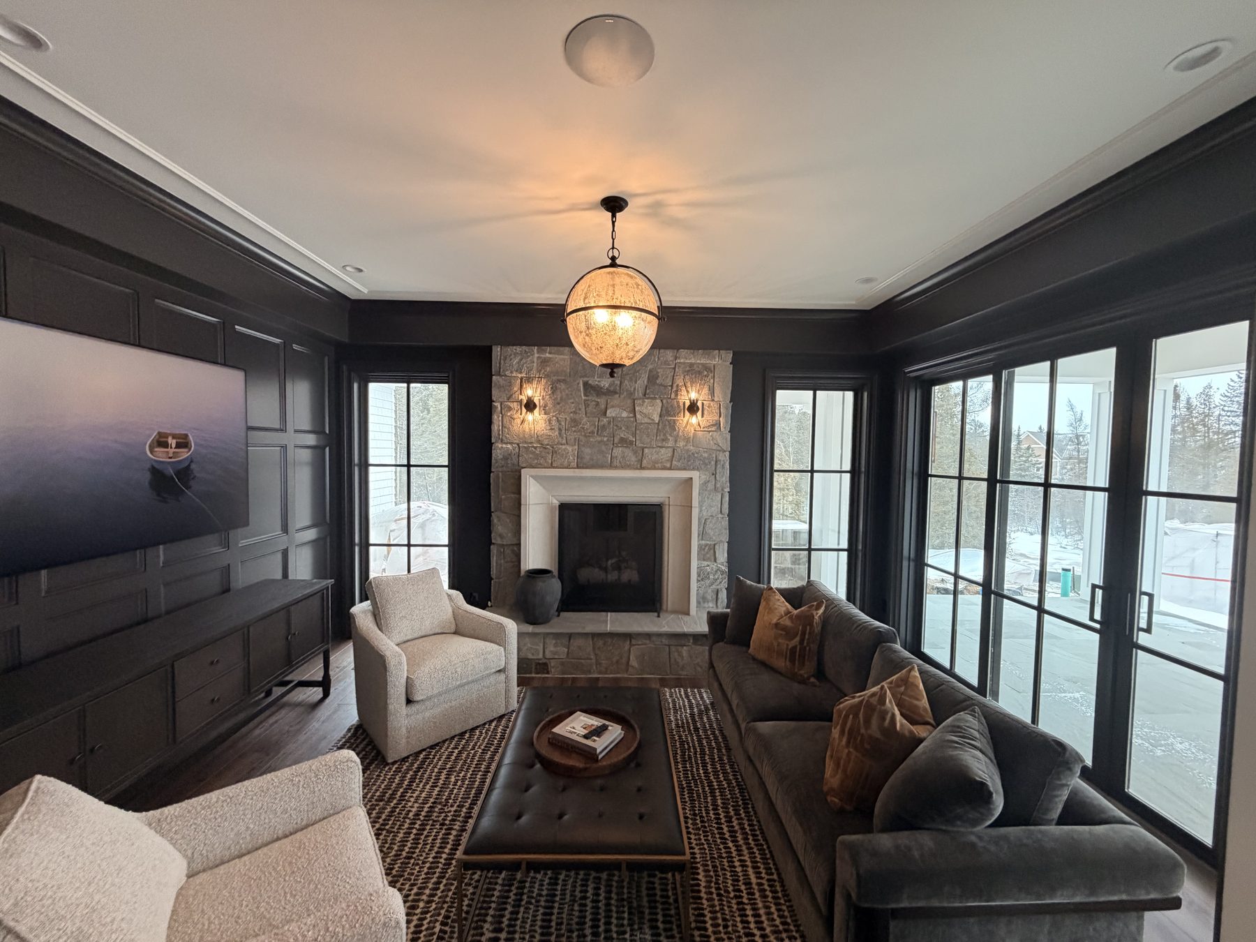Living room with a large wall-mounted TV in a dark coffered wall to the left, a stone fireplace with paired sconces in the middle, gray velvet sofa, ottoman, and large windows overlooking a snowy landscape