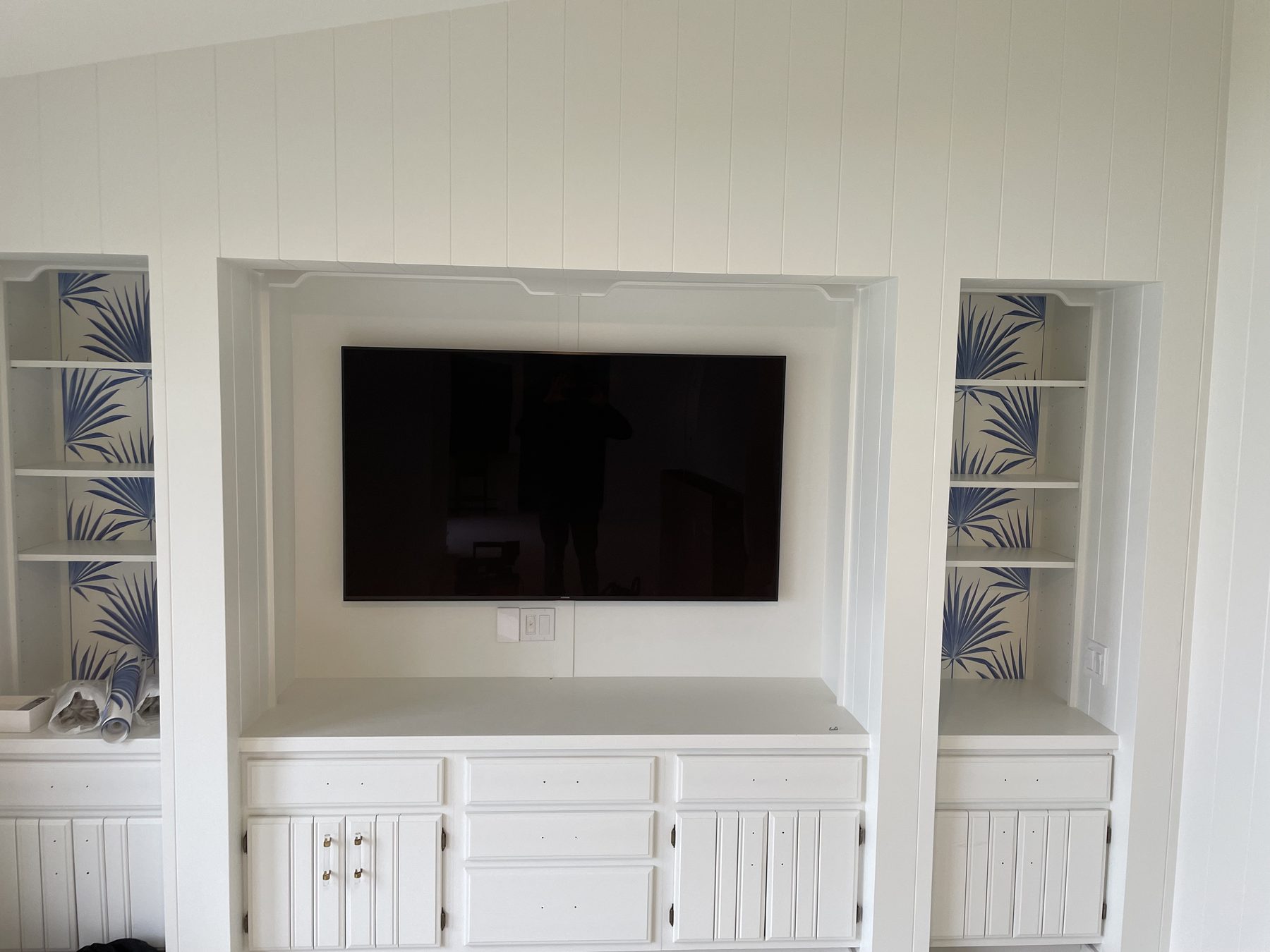 TV flush-mounted in a white shiplap millwork niche flanked by display shelves with palm-leaf wallpaper